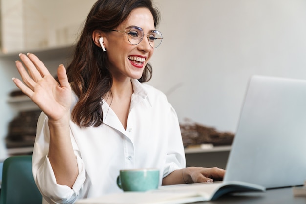 Aantrekkelijke vrolijke jonge brunette zakenvrouw zittend aan de cafétafel met laptopcomputer binnenshuis, het dragen van draadloze oortelefoons, audio-oproep, zwaaiende hand