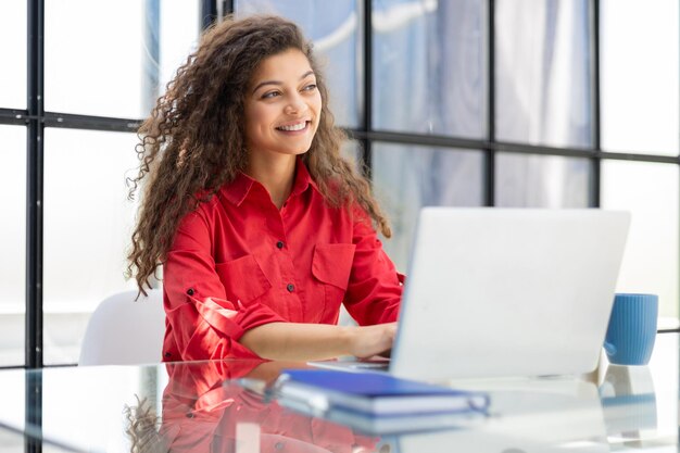 Aantrekkelijke vrolijke zakenvrouw in rood shirt die op laptop werkt op modern kantoor