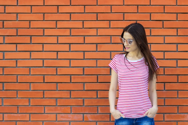 Aantrekkelijke vrouw die gestreepte t-shirt en modieuze zonnebril dragen die tegen rode bakstenen muur stellen, swag straatstijl. Leeg