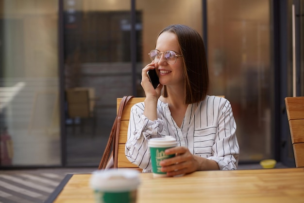 Aantrekkelijke vrouw die op mobiele telefoon praat terwijl ze in de coffeeshop zit en warme drank drinkt terwijl ze een gesprek heeft met een smartphone terwijl ze pauze heeft in het café