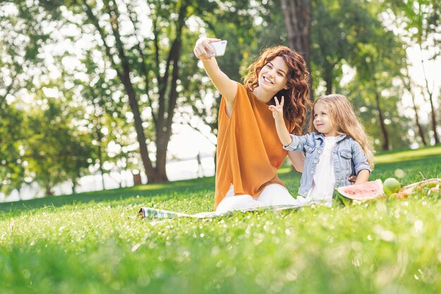 Aantrekkelijke vrouw en meisje nemen selfie in het park zittend op de picknickdeken