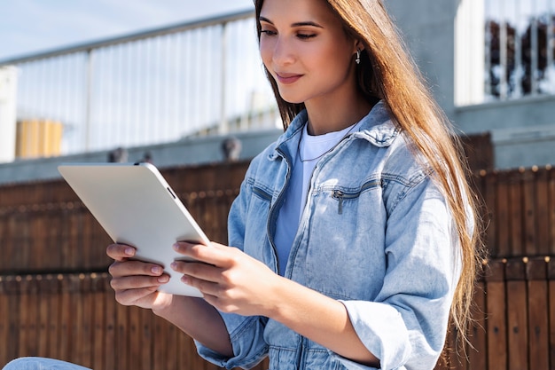 Aantrekkelijke vrouw in denim kleding zit op een bankje, houdt slimme tablet in haar handen. portret van mooie vrouw