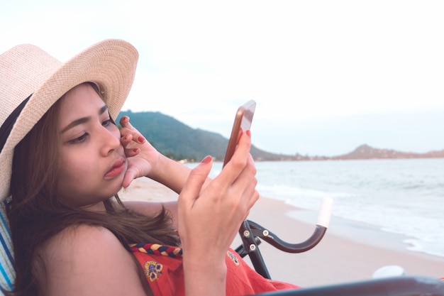 Aantrekkelijke vrouw in hoed die smartphone gebruiken terwijl het zitten op ligstoel bij zonsondergangstrand.