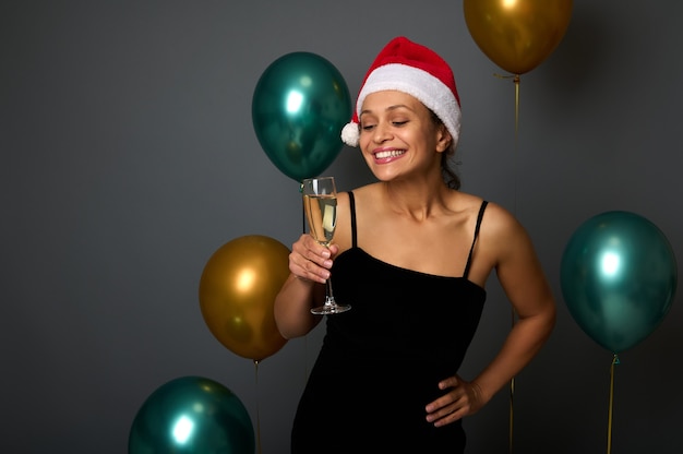 Aantrekkelijke vrouw in kerstmuts en zwarte jurk heeft plezier op kerstfeest, glimlacht brede glimlach kijken camera met champagne fluit, geïsoleerd op een grijze achtergrond met glanzende gouden groene lucht ballonnen