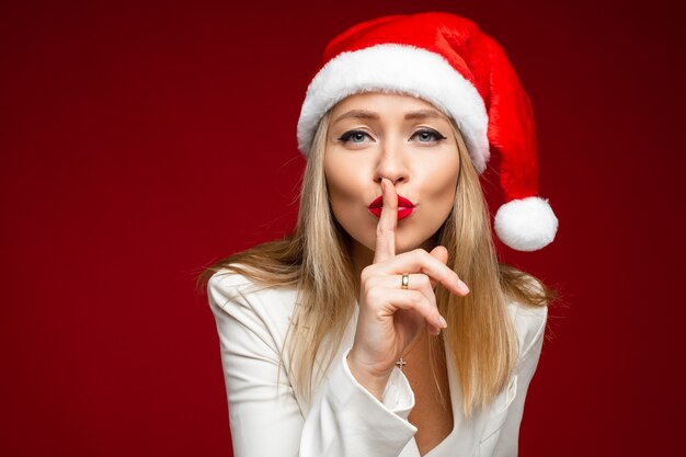 aantrekkelijke vrouw in kerstmuts met wijsvinger op lippen vragen stil te zijn. Geheim of stilteconcept isoleren op rode muur.