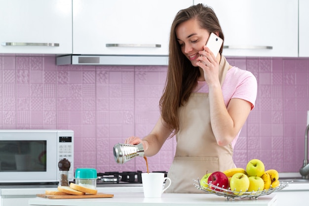 Aantrekkelijke vrouw in schort die op telefoon spreekt en hete Turkse ochtendkoffie van cezve voor ontbijt bij keuken maakt