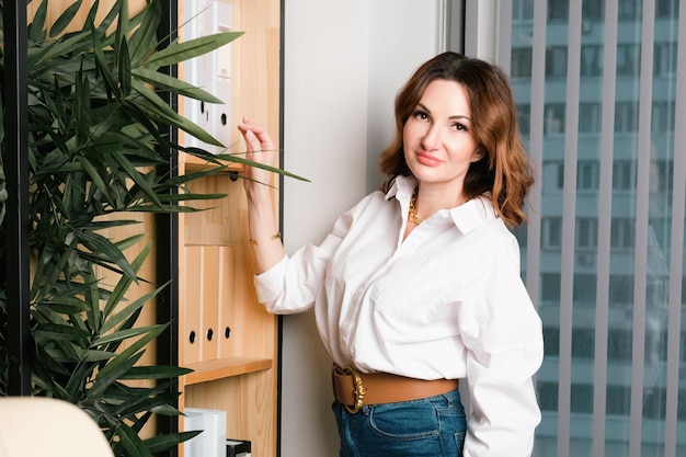 Aantrekkelijke vrouw in spijkerbroek en witte blouse haalt map met documenten uit een plank