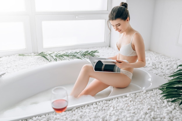 Aantrekkelijke vrouw in wit ondergoed tijdschrift lezen en ontspannen in bad, glas rode wijn staande op de rand. luxe badkamer interieur, stenen decor