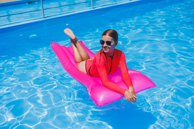 Aantrekkelijke vrouw in zonnebril is ontspannen en zonnebaden op een roze matras in het zwembad Jonge vrouw in een roze zwempak drijft op een opblaasbare roze matras