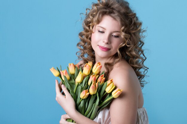Aantrekkelijke vrouw met blond krullend haar boeket bloemen houden over blauwe achtergrond.