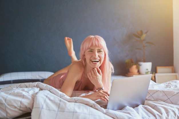 Aantrekkelijke vrouw met felroze lacht met behulp van moderne laptop op bed bij blauwe muur