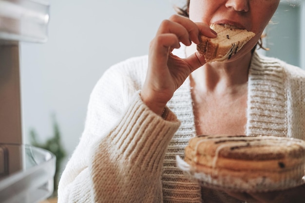 Aantrekkelijke vrouw van middelbare leeftijd in een gezellig vest die cake uit de koelkast eet in de keuken thuis