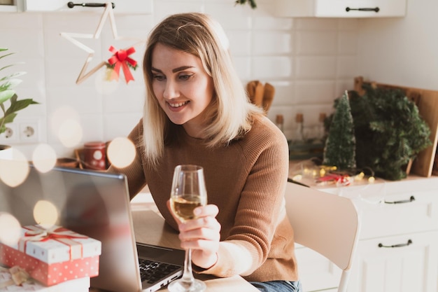 Aantrekkelijke vrouw videochatten voor kerstvakantie in de keuken met glas champagne en geschenken