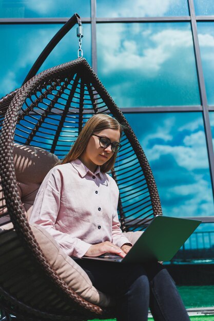 Aantrekkelijke vrouw zit met een laptop op het balkon en kijkt aandachtig naar het scherm Een mooi freelancer-meisje in vrijetijdskleding werkt op een laptop terwijl ze in een stoel op het balkon zit