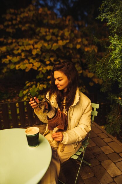 aantrekkelijke vrouw zit 's avonds in een café thee te drinken met een hond en gebruikt een smartphone