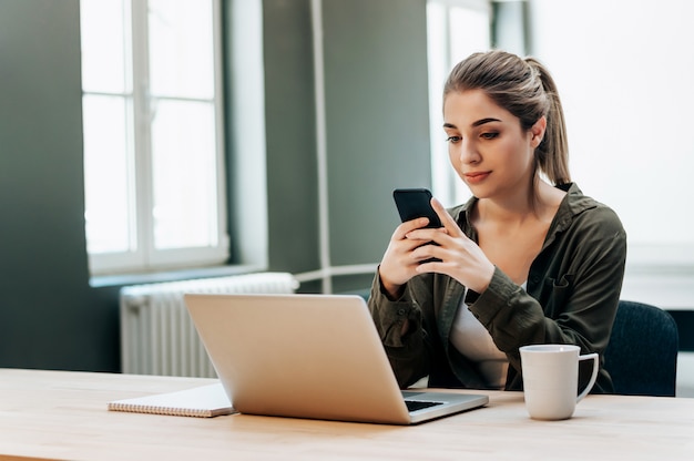 Aantrekkelijke vrouwelijke freelancer houd slimme telefoon terwijl het zitten voor laptop.