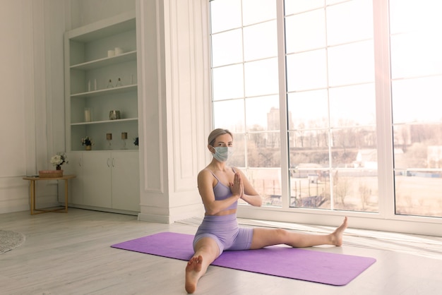 Aantrekkelijke vrouwelijke sportfitnesscoach die een wit medisch masker draagt, doet thuis oefeningen op yogamat.