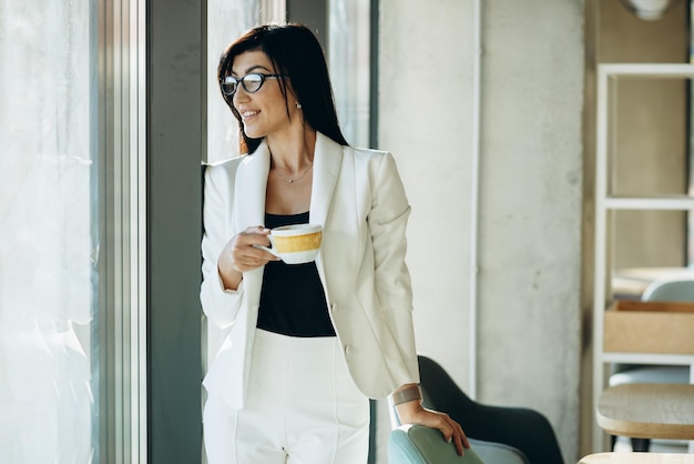 Aantrekkelijke zakenvrouw die koffie drinkt op kantoor bij het raam