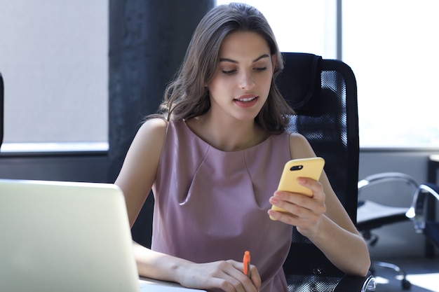 Aantrekkelijke zakenvrouw met behulp van slimme telefoon zittend aan het bureau.