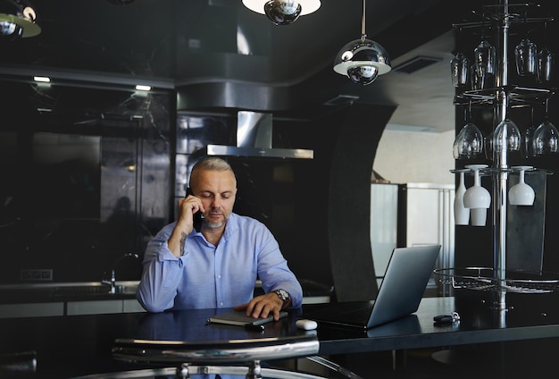 Aantrekkelijke zelfverzekerde freelancer, succesvolle zakenman die op mobiele telefoon praat terwijl hij op afstand werkt op een laptop in zijn stijlvolle studiokeuken