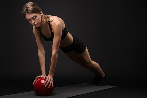 Aantrekkelijke zelfverzekerde jonge gezonde fitnessvrouw die sportbeha en korte broek draagt, geïsoleerd op zwarte achtergrond, traint op een fitnessmat en medicijnbal gebruikt