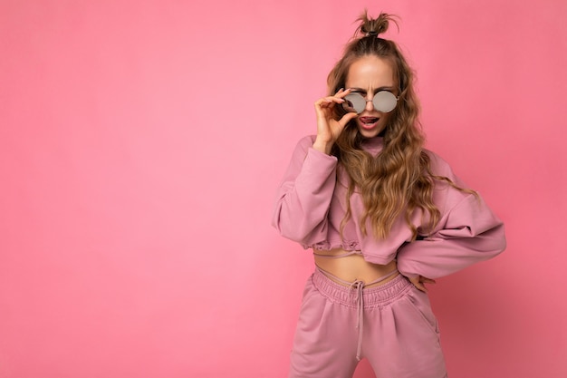 Aantrekkelijke zelfverzekerde positieve jonge blonde vrouw die alledaagse stijlvolle kleding en moderne zonnebril draagt, geïsoleerd op een kleurrijke achtergrondmuur die naar de camera kijkt