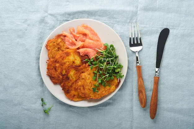 Aardappelpannenkoekjes Gebakken zelfgemaakte aardappelpannenkoekjes of latkes met crème groene uien microgreens rode zalm en saus in rustieke plaat op blauw linnen tafelkleed Rustieke stijl Gezonde voeding Bovenaanzicht