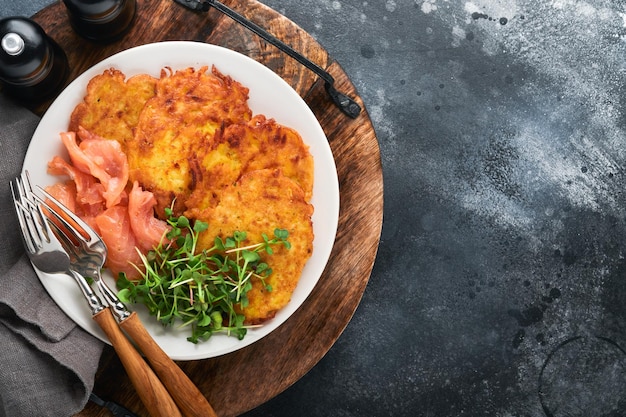 Aardappelpannenkoekjes Gebakken zelfgemaakte aardappelpannenkoekjes of latkes met crème groene uien microgreens rode zalm en saus in rustieke plaat op oude zwarte tafel achtergrond Rustieke stijl Bovenaanzicht