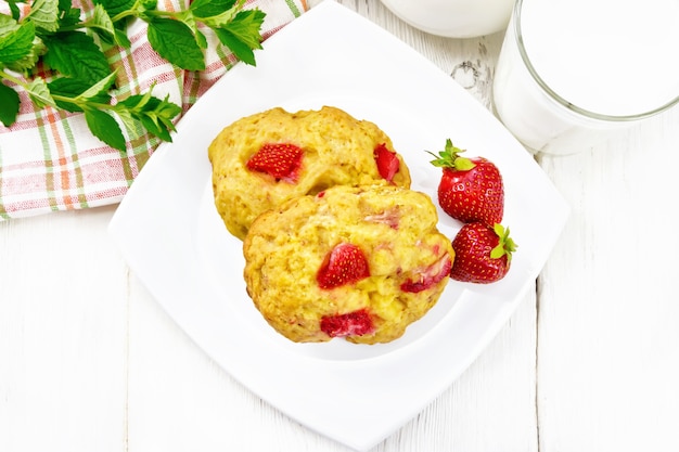 Aardbeienscones in een bord met bessen, een servet, munt, melk in kruik en glas op de achtergrond van houten plank van bovenaf