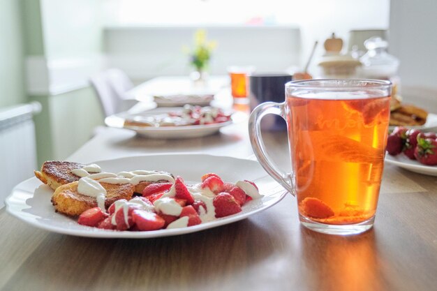 Aardbeienseizoen, eten en drinken met bessen. Eten op tafel, wrongelpannenkoekjes met aardbeien en zure room. Thee met citroen en bessen