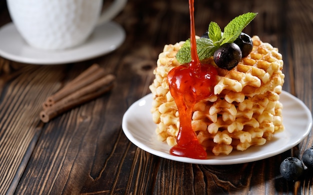 Foto aardbeiensiroop gieten op een stapel versgebakken belgische wafels versierd met bosbessen en munt op witte plaat heerlijk zelfgemaakt gebak voor het ontbijt op houten tafel of rustieke achtergrond