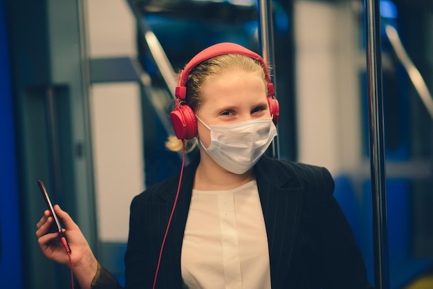 Aardig, behoorlijk bezorgd jong meisje met een masker en koptelefoon in een bustrein of metro die naar school gaat