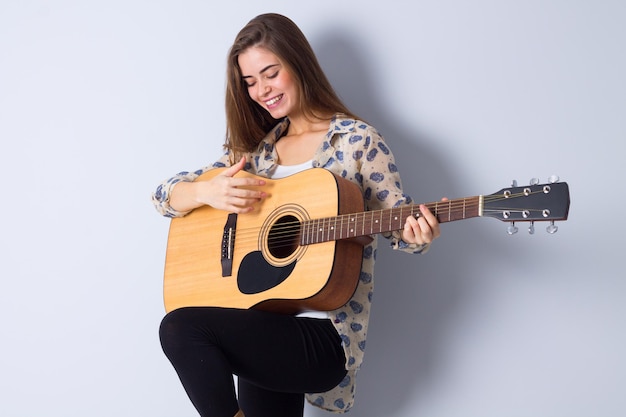 Aardige jonge vrouw met lang haar in bruine jas gitaar spelen op witte achtergrond in studio