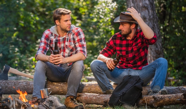 Aardige man drinkt bier bij vreugdevuur ranger bij buitenactiviteiten picknick weekend doorbrengen in de natuur avontuur concept wandeling en mensen twee mannen ontspannen bij vuur wandelen en kamperen mannelijke vriendschap