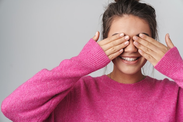 aardige opgewonden vrouw in roze trui glimlachend en bedekt haar ogen geïsoleerd over een witte muur