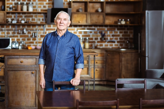 Aardige oude man die in de keuken staat terwijl hij op de stoel leunt