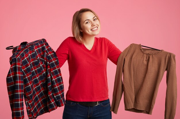 Foto aarzelend mooie jonge vrouw houdt twee shirts in handen, kan niet kiezen welke te kopen, ziet er positief uit