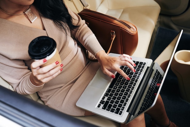 Фото Выше вид неузнаваемой бизнес-леди с одноразовой чашкой кофе, печатающей на ноутбуке в такси