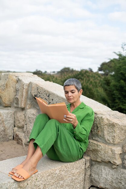 公園で読書をしている年配の女性に夢中。本を保持している明るい服を着た短い白髪の女性モデル。レジャー、文学の概念