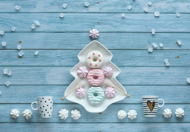 Photo abstract decorative christmas background with sweets, coffee cups, marshmallows, doughnuts and sugar crystals. plate in xmas tree shape and sweets arranged on rustic blue wooden background.