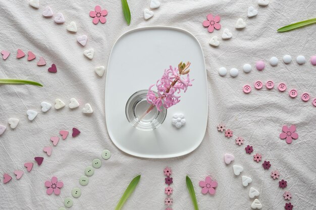 Abstract Springtime background Pink pearl hyacinth flower in glass on ceramic plate Flat lay with hearts candy buttons small decor arranged on white textile background