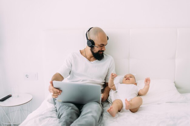 An abusive man with a beard in a shirt and jeans sits on the bed with a laptop in his hands and his little child lies next to him