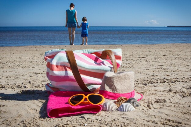ビーチの砂でリラックスするためのアクセサリーと海の背景にある母親と息子の旅行と休暇の時間