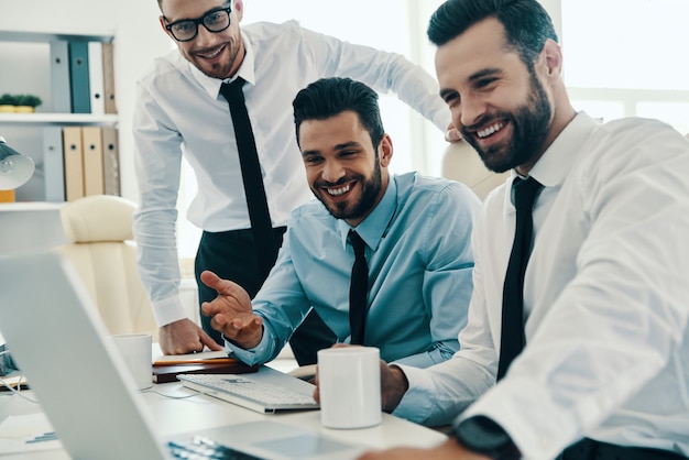 Achieving best results together. Group of young modern men in formalwear working using computers and smiling while sitting in the office