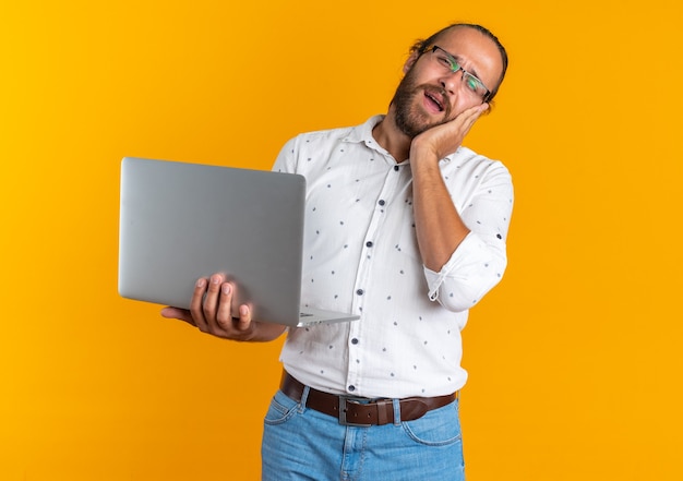 Aching adult handsome man wearing glasses holding laptop keeping hand on face looking at camera isolated on orange wall