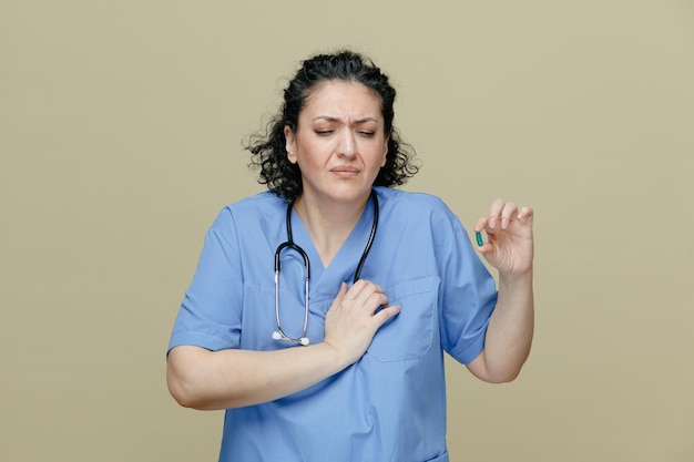 Aching middleaged female doctor wearing uniform and stethoscope around neck showing capsule keeping hand on chest with closed eyes having heartache isolated on olive background