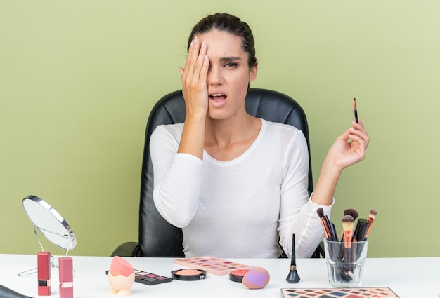 Aching pretty caucasian woman sitting at table with makeup tools putting hand on her eye and holding makeup brush isolated on olive green wall with copy space