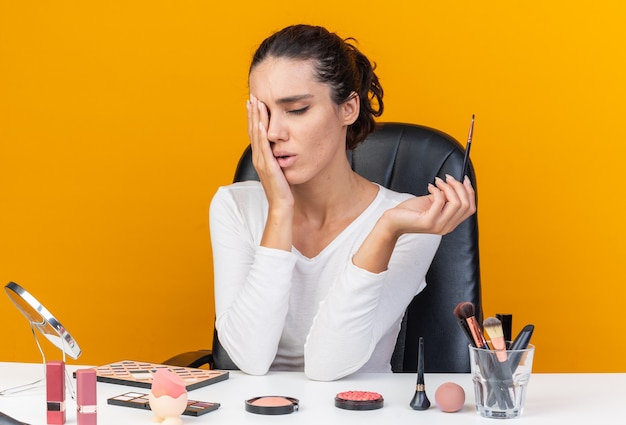 Aching pretty caucasian woman sitting at table with makeup tools putting hand on her eye and holding makeup brush isolated on orange wall with copy space