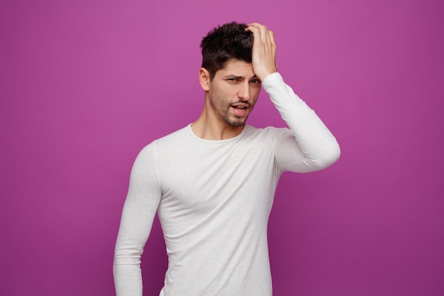 Aching young handsome man looking at camera keeping hand on head on purple background