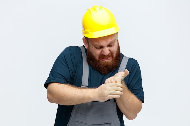 Aching young male construction worker wearing safety helmet and uniform holding fingers looking at them isolated on white background